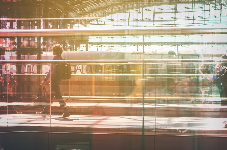 Ein junger Mann mit Rucksack und einer Tasche geht über ein Gleis in einem Bahnhof. Im Hintergrund sind ein Zug, möglicherweise ein ICE, und das Bahnhofsgebäude zu sehen. Das Bild ist durch eine verschwommen reflektierende Scheibe aufgenommen.