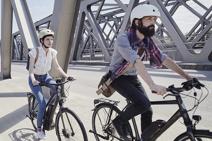 Das Bild zeigt im Bildmittelpunkt sind eine Frau und ein Mann auf Fahrrädern zu sehen, die über eine Brücke fahren. Daneben steht ein Willkommenstext auf blauem Hintergrund.